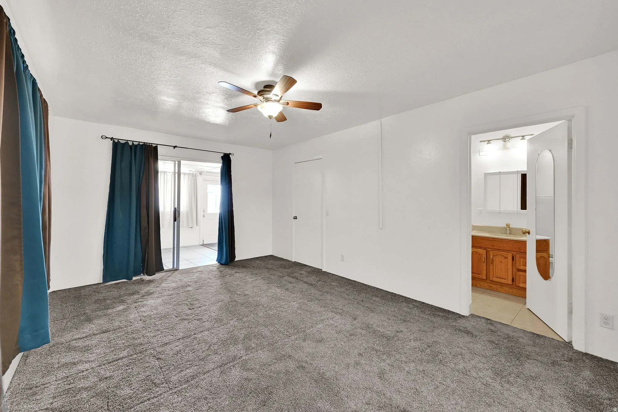 Carpeted room with a ceiling fan, sliding glass door access to a tiled outdoor space, and an adjacent bathroom featuring a wood vanity and wall-mounted light fixture