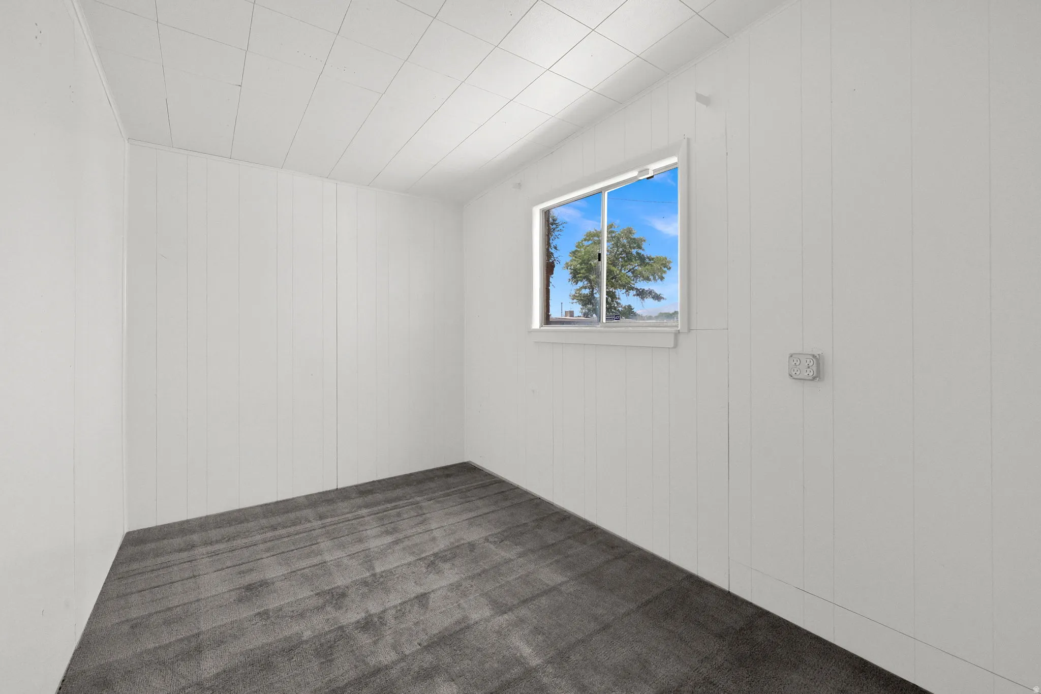 Room featuring white vertical paneling, gray patterned carpeting, a single window, and a white grid ceiling