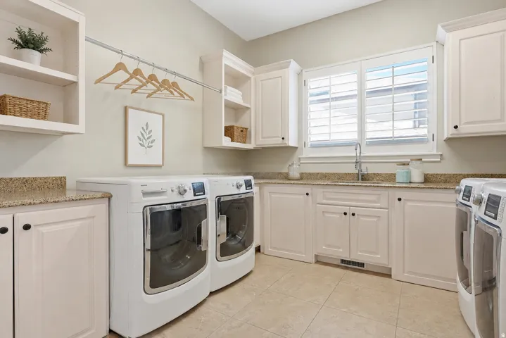 Functional utility room featuring extensive white cabinetry, granite-finish countertops, a built-in sink with a gooseneck faucet, tiled flooring, and a window with white plantation shutters