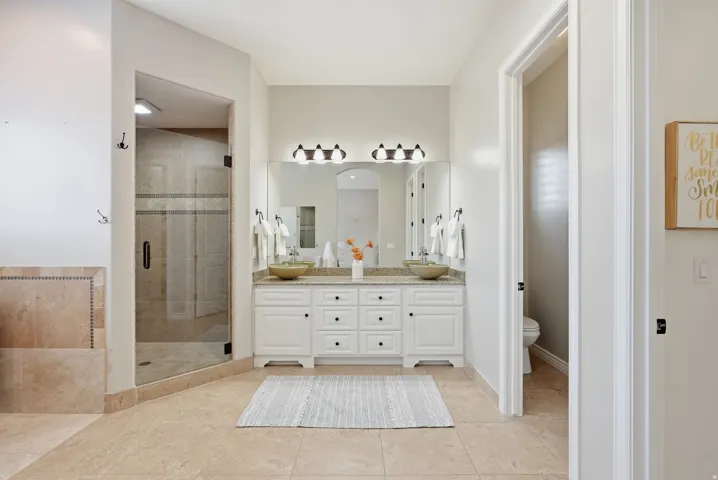 Dual vessel sinks set in a white vanity with a light-toned countertop, under a large mirror and three-bulb vanity light