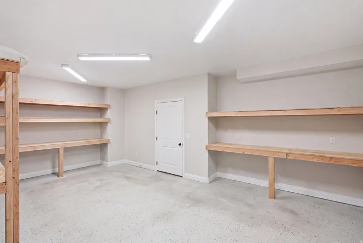 Spacious utility room featuring extensive built-in wooden shelving, a sturdy built-in workbench, durable epoxy flooring, bright overhead fluorescent lighting, and a solid panel door