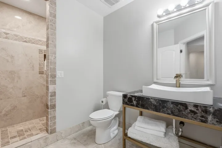 Bathroom featuring a walk-in shower with tile surround, a dark stone-finish vanity top, a vessel sink with a gold-tone faucet, and a wall-mounted mirror with overhead lighting