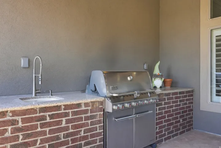 Outdoor kitchen featuring a built-in stainless steel grill, integrated sink with a gooseneck faucet, brick base cabinetry, and a light-toned countertop