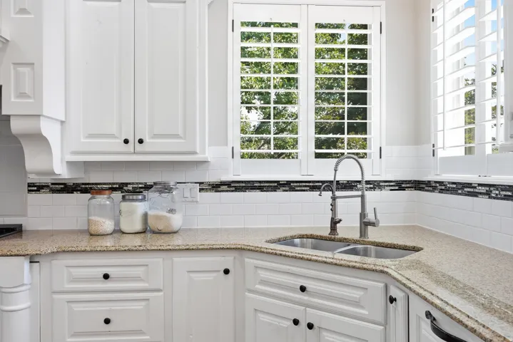 Kitchen featuring white cabinetry, light-toned countertops, a double basin sink with a pull-down faucet, and white subway tile backsplash with a decorative mosaic accent