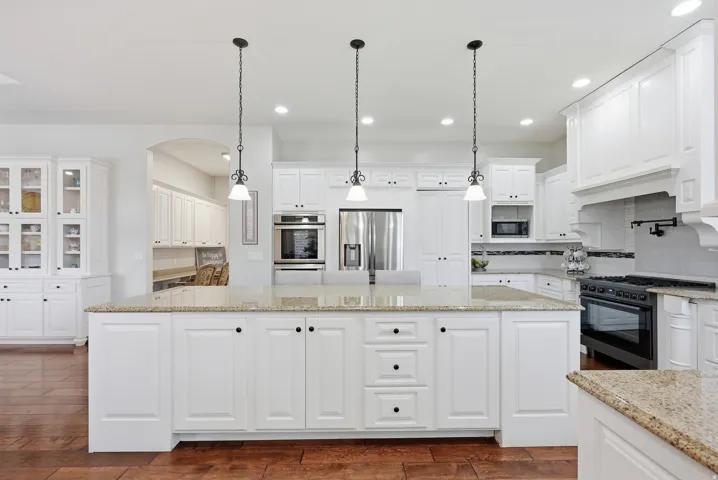 Spacious kitchen featuring a large central island with light-toned stone countertops, abundant white cabinetry, integrated stainless steel appliances, and wood-finish flooring