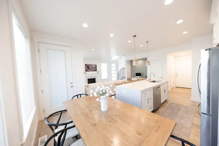 Open-concept kitchen and dining area featuring a light-toned wood dining table, white kitchen island with an undermount sink, stainless steel dishwasher, and recessed lighting throughout