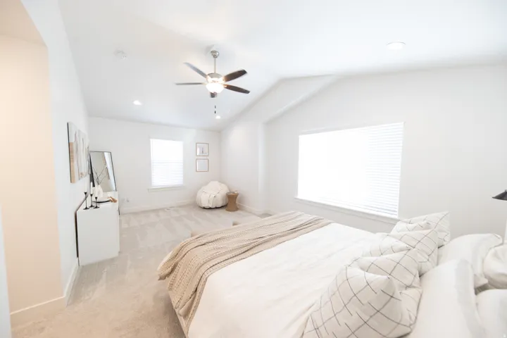 Vaulted ceiling bedroom with light gray carpeting, featuring a ceiling fan with integrated lighting, two windows with blinds, and recessed lighting fixtures