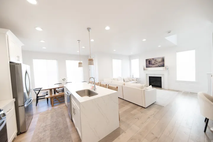 Open-concept living space featuring wood-finish flooring, a kitchen island with a stainless steel sink, recessed lighting, a fireplace with a mantel, and multiple windows