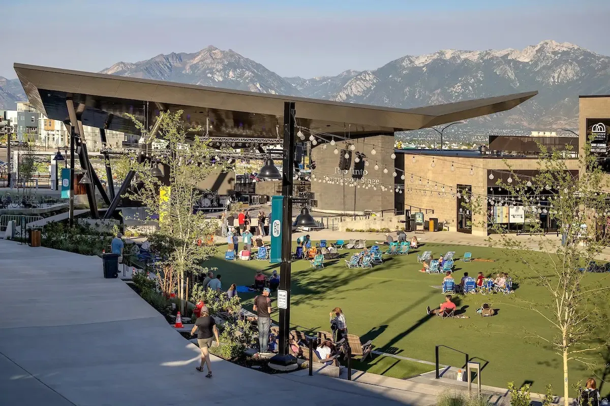 Central green space featuring a performance stage with a striking architectural canopy, surrounded by commercial buildings with brick and light-colored facades