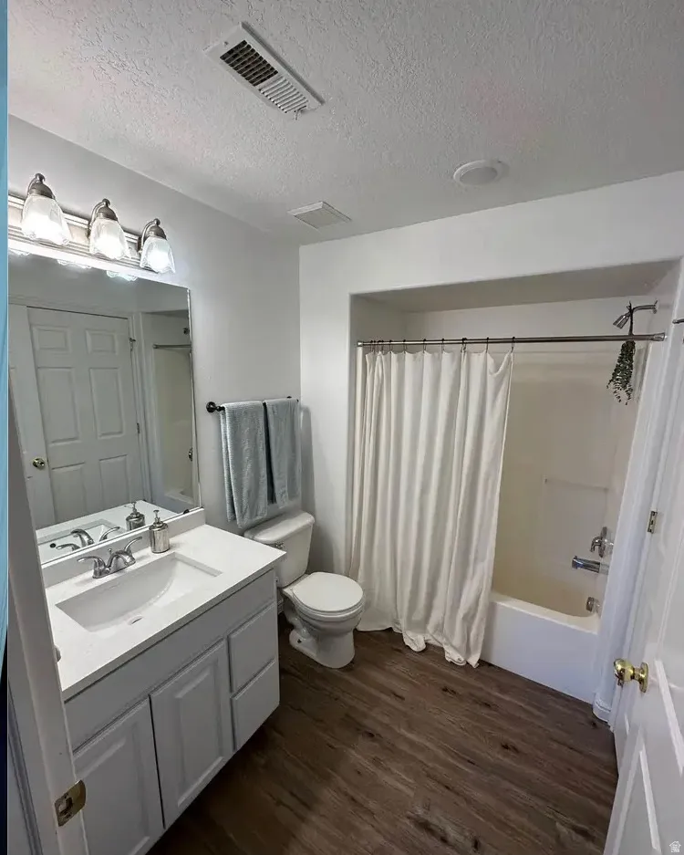 Bathroom featuring a white vanity with an integrated sink and chrome fixtures, wood-finish flooring, a full-sized mirror, a white toilet, and a shower-tub combination