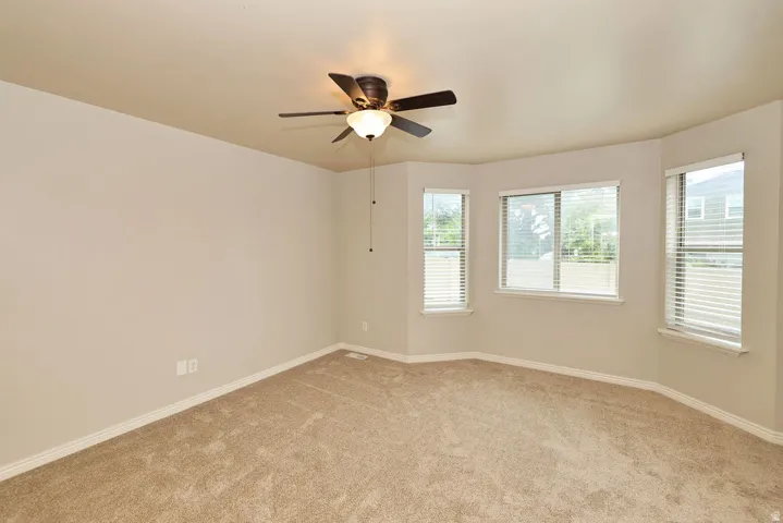 Bright corner bedroom featuring neutral carpeting, ceiling fan with integrated lighting, and abundant natural light from multiple windows with blinds