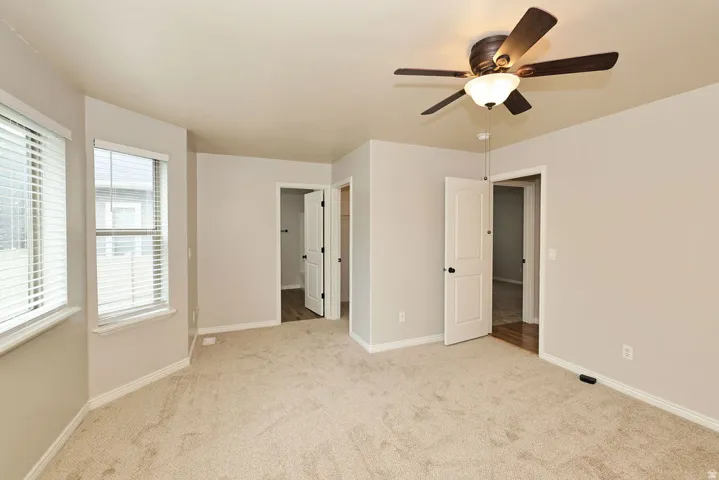 Spacious primary bedroom featuring neutral carpeting, abundant natural light from dual windows with plantation shutters, ceiling fan with integrated lighting, and direct access to ensuite and walk-in closet