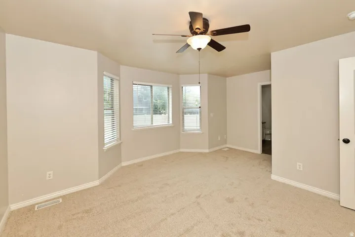 Spacious bedroom featuring neutral carpet flooring, multiple windows with plantation shutters providing natural light, ceiling fan with integrated lighting fixture, and white six-panel doors