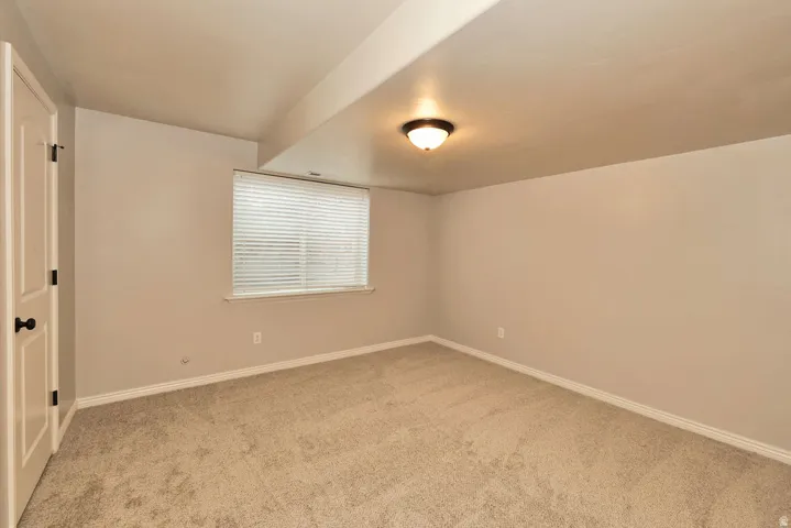 Neutral-toned room featuring a window with blinds, recessed ceiling beam, flush-mount lighting, and light-colored carpet