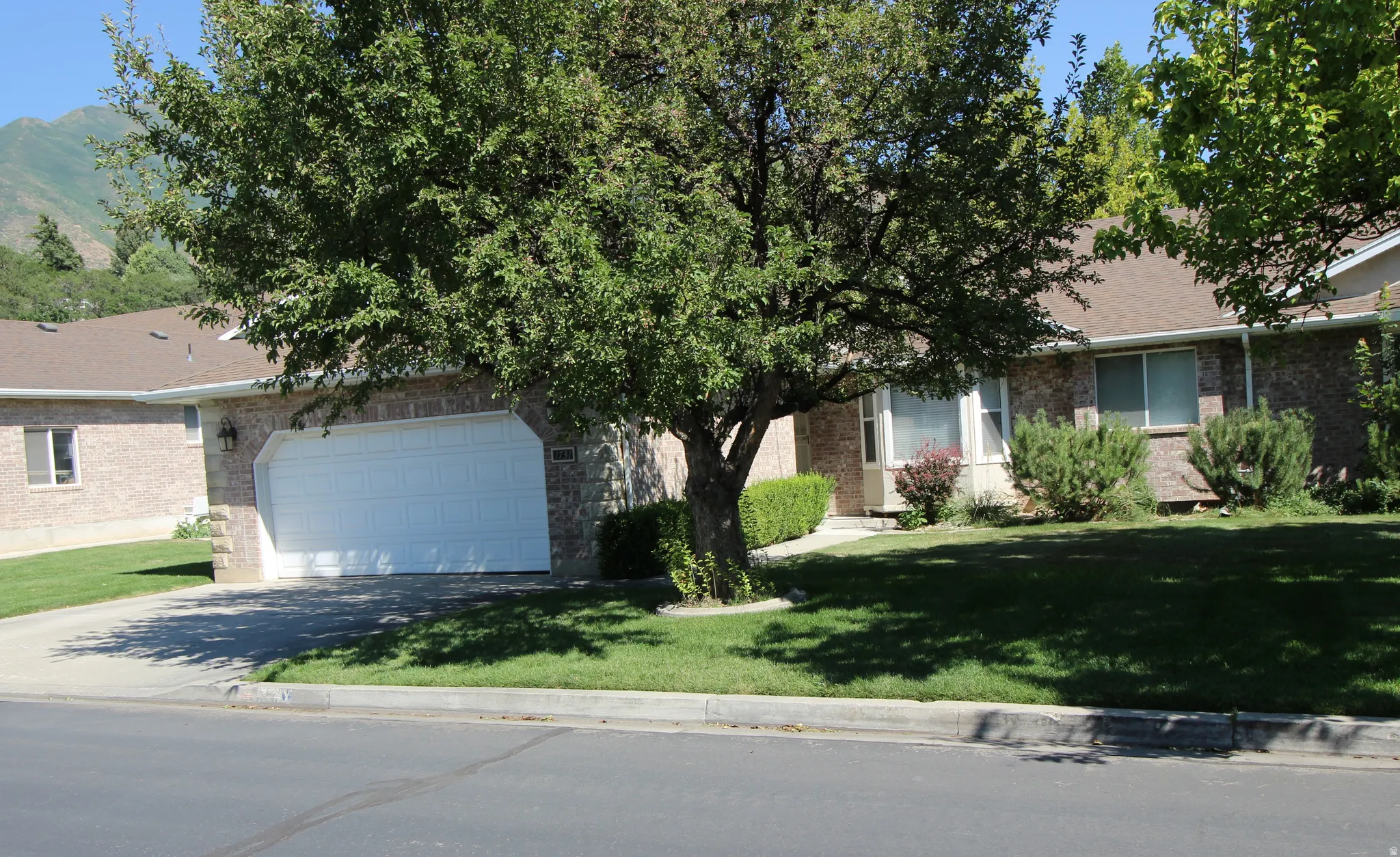 Brick exterior featuring a two-car garage, mature trees, and a mountain backdrop