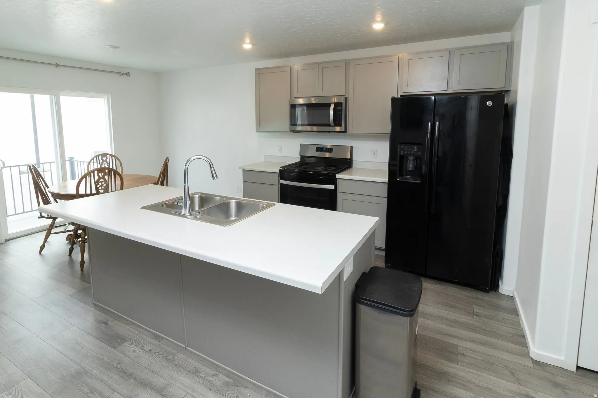 Modern kitchen featuring an island with an integrated double basin sink, light gray cabinetry, white countertops, black appliance suite, and wood-finish flooring