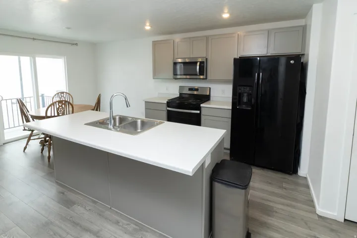 Modern kitchen featuring an island with an integrated double basin sink, light gray cabinetry, white countertops, black appliance suite, and wood-finish flooring