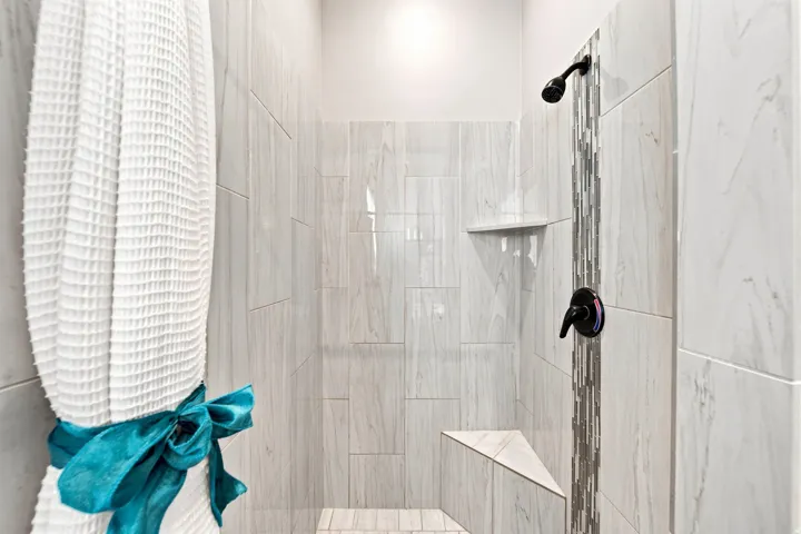 Tiled shower enclosure featuring light-toned large format tiles, a decorative vertical mosaic accent, black shower fixtures, a built-in shelf, and a corner shower bench