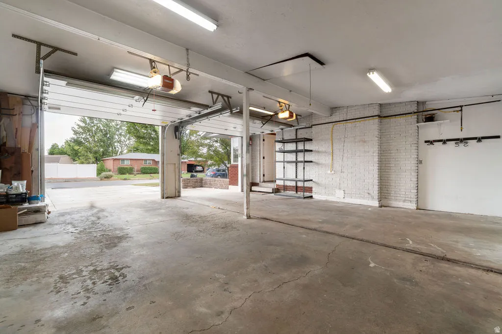 Spacious two-car garage featuring a concrete floor, white painted brick wall, built-in shelving, and exposed fluorescent lighting