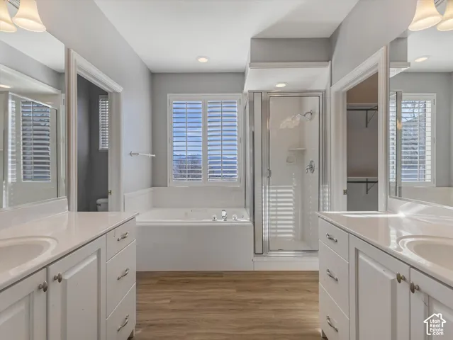 ensuite Bathroom featuring dual vanities with white cabinetry and integrated sinks, a built-in jetted tub under a window with plantation shutters, a glass-enclosed shower, and wood-finish flooring