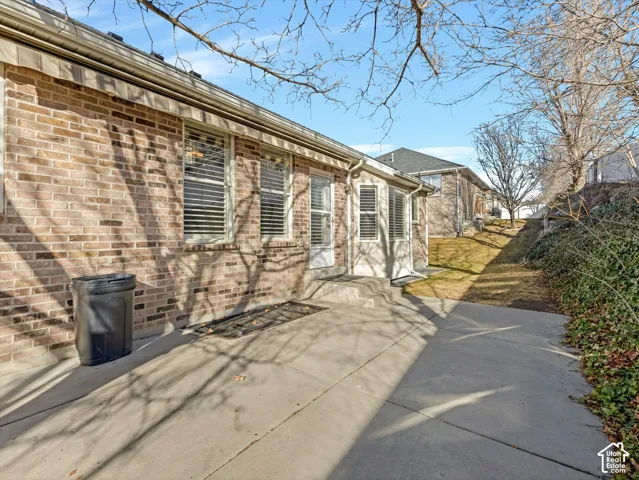 Concrete patio with a brick exterior, multiple windows with blinds, and a sloped, landscaped yard