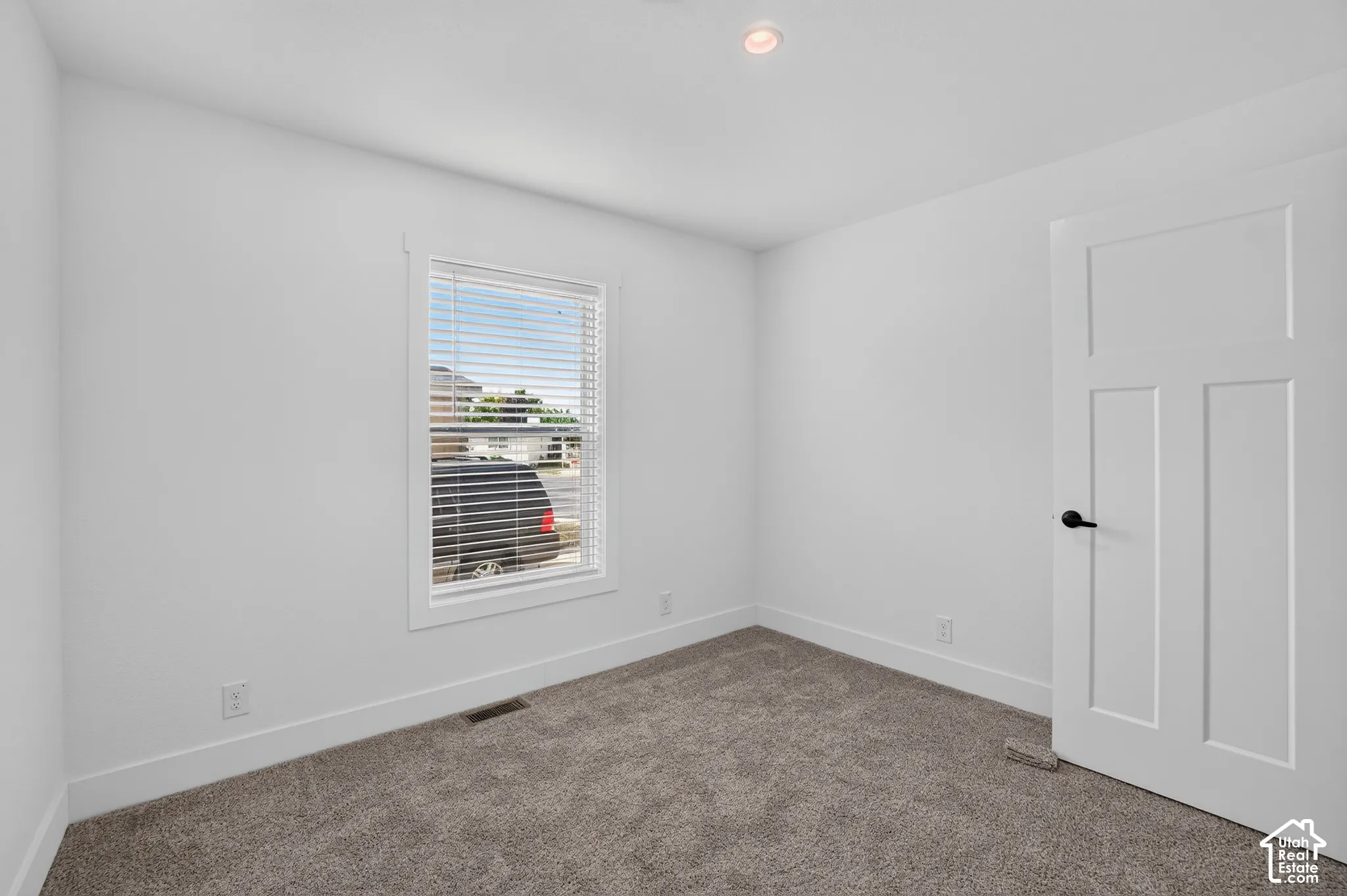 Carpeted room with a single window featuring blinds, a white paneled door with matte black hardware, recessed ceiling lighting, and crisp white trim