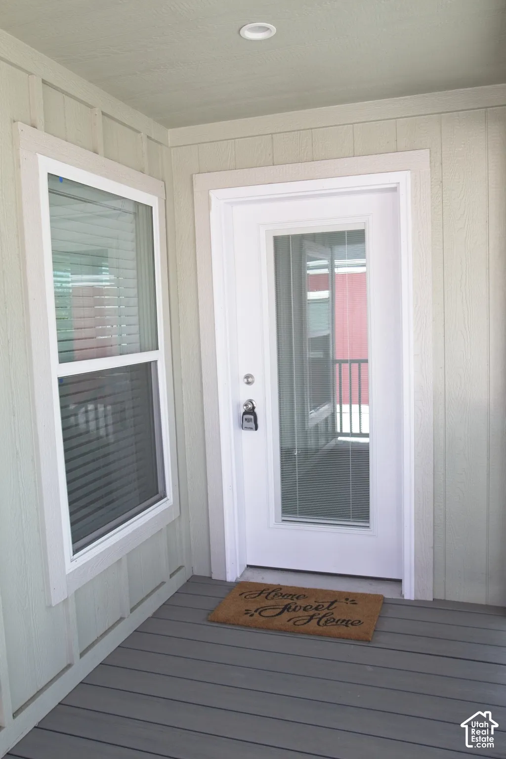 Entryway featuring vertical board-and-batten siding, a white door with a glass lite, a single-hung window with integrated blinds, and a covered porch with gray composite decking