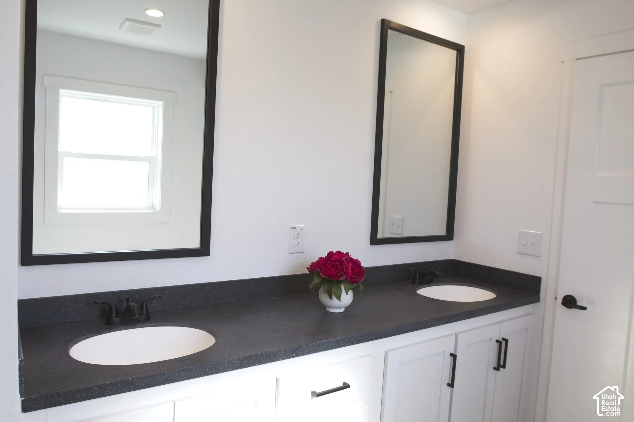 Double vanity featuring a dark solid-surface countertop, dual drop-in sinks, matte black fixtures, and framed mirrors, finished with white shaker-style cabinetry and matte black hardware