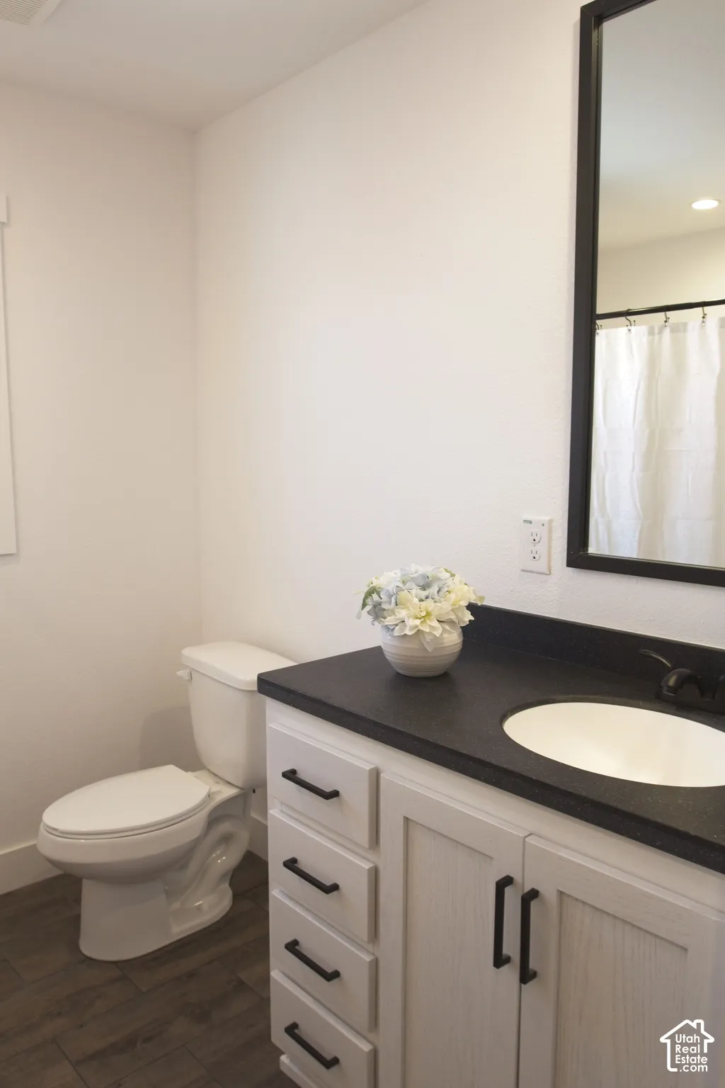 White vanity featuring a dark countertop, integrated oval sink, matte black hardware, wood-finish flooring, and a framed mirror