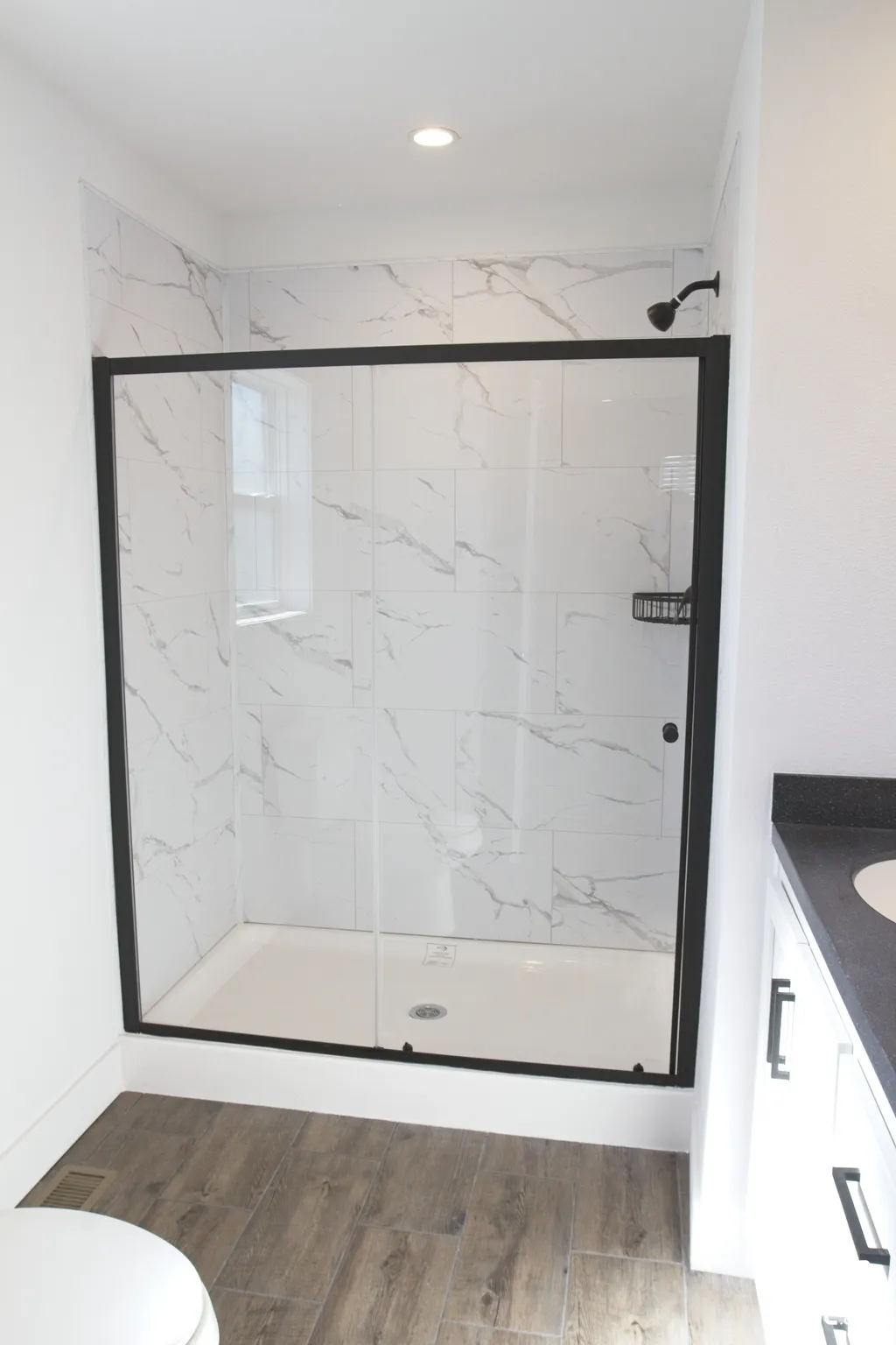 Walk-in shower featuring white tile with grey veining, a matte black sliding glass enclosure, a fixed shower head, and wood-finish flooring