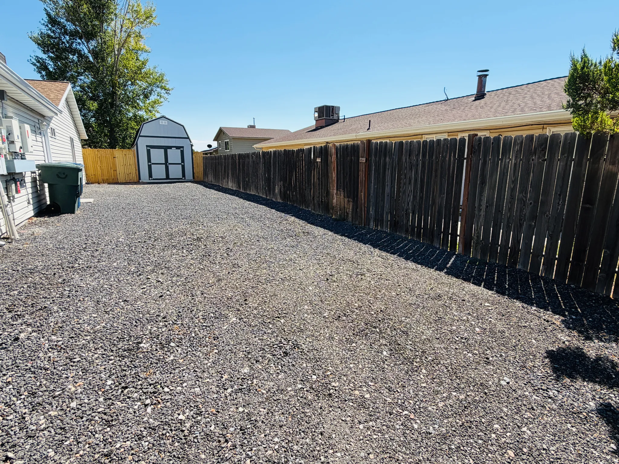 Gravel yard featuring a gray storage shed with white trim and double doors, wood fencing, and an exterior wall with utility connections