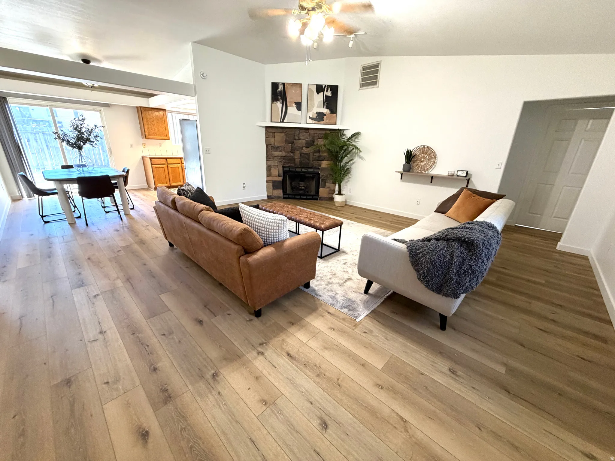 Open-concept living area featuring light wood-finish flooring, a stone-clad fireplace with mantel, and a vaulted ceiling with a ceiling fan