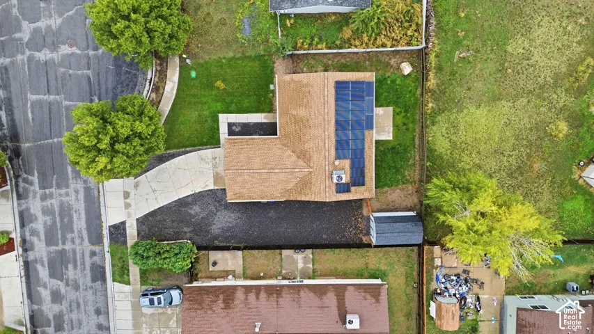 Aerial perspective of a residential lot featuring a concrete driveway, roof-mounted solar panels, and a fenced yard with mature trees