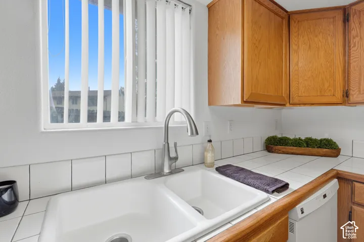 Double-basin sink with a high-arch gooseneck faucet, tile countertops, square tile backsplash, and wood-finish cabinetry