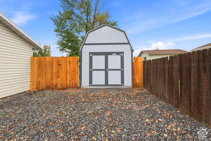 Detached storage structure featuring a gambrel roof design, dual-door entry, and gravel ground cover, enclosed by wood privacy fencing