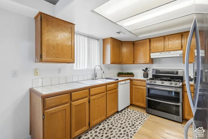 Kitchen featuring wood cabinetry, tile countertops, stainless steel gas range, white double-basin sink, and wood-finish flooring