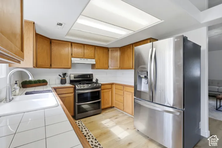 Kitchen featuring wood cabinetry, white tiled countertops, stainless steel appliances, a double-basin sink, and wood-finish flooring