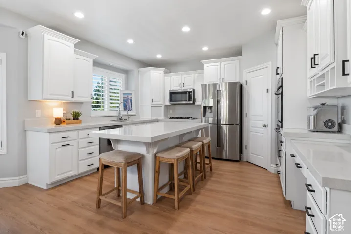 Kitchen featuring white cabinetry with black hardware, light-toned countertops, a central island with seating, stainless steel appliances, and wood-finish flooring