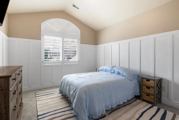 Vaulted ceiling bedroom featuring board and batten wainscoting, an arched window with louvered shutters, and light-colored carpeting