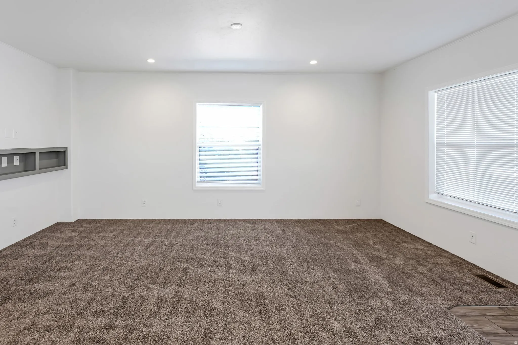 Spacious room with plush brown carpeting, recessed lighting, and two windows featuring horizontal blinds