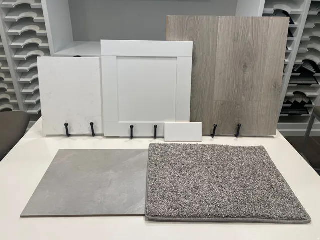 Modern interior finishes featuring a white shaker cabinet door, light wood-finish flooring, a grey large-format tile, a textured grey carpet sample, and a white countertop material