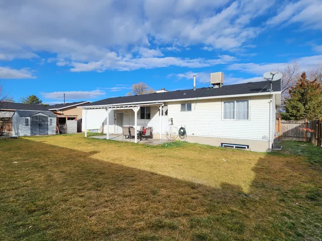 Expansive grass lawn featuring a covered patio, a detached storage shed, white vinyl siding, a black shingle roof, and a wood privacy fence