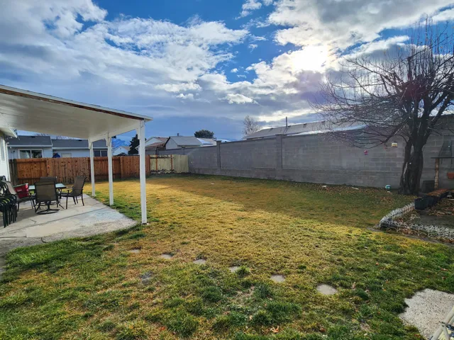 Expansive backyard featuring a covered patio with concrete flooring, a large lawn area, a mature tree, and perimeter fencing composed of wood and concrete block