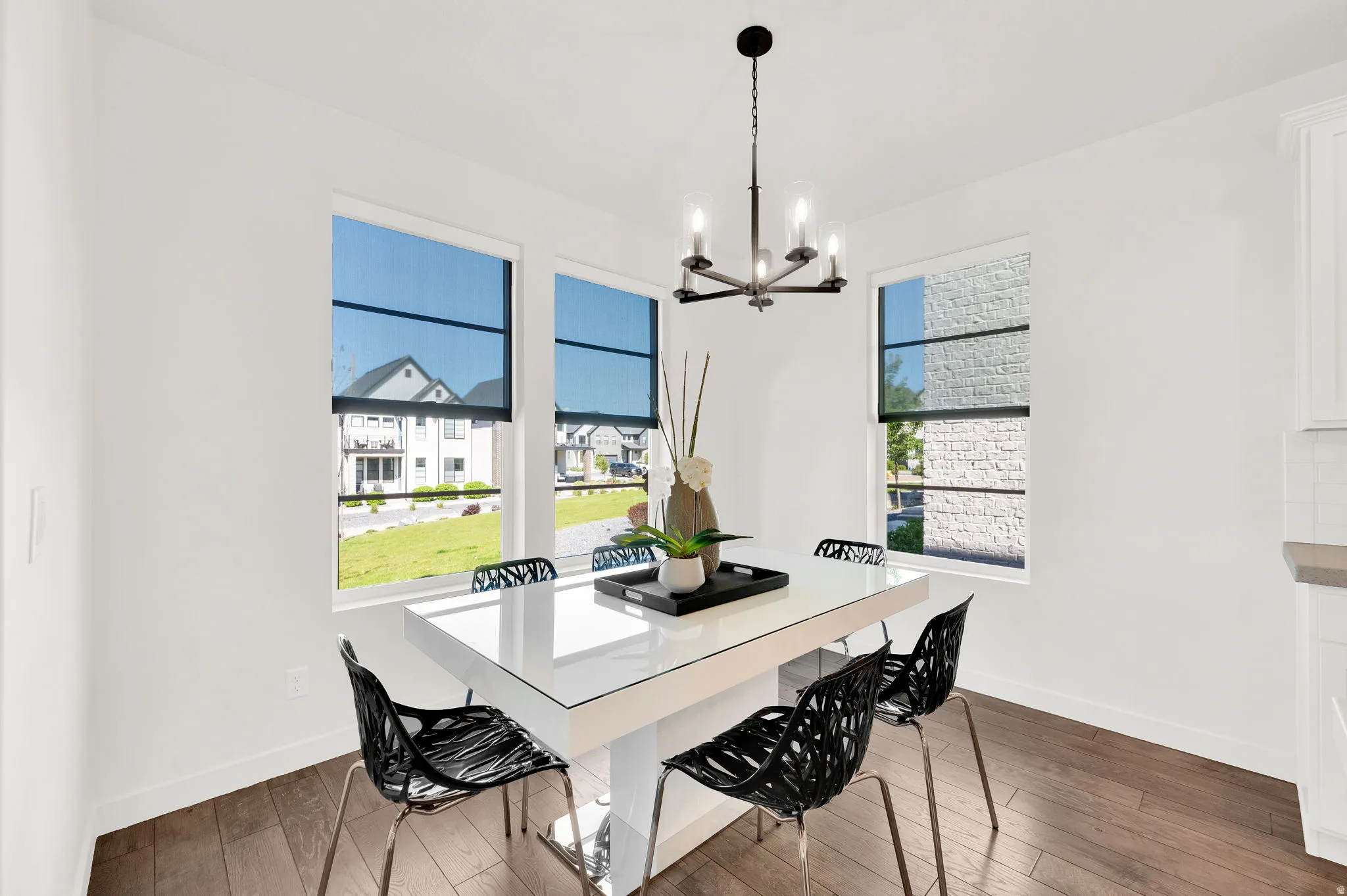 Bright dining area featuring a modern chandelier, dark wood-finish flooring, and multiple windows with dark roller shades