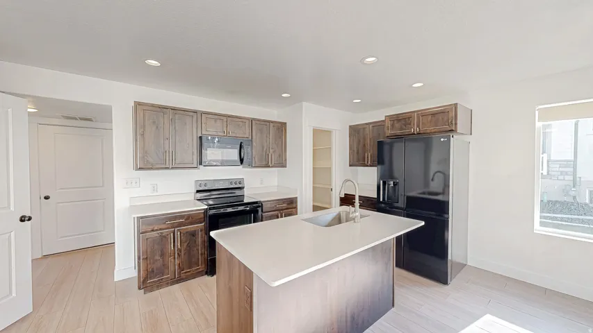 Spacious kitchen featuring an island with an undermount sink, light-toned wood-finish flooring, recessed lighting, wood cabinetry, and a full suite of black appliances