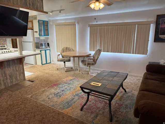 Living area featuring carpet flooring, vertical blinds on a large window, and a ceiling fan with integrated lighting