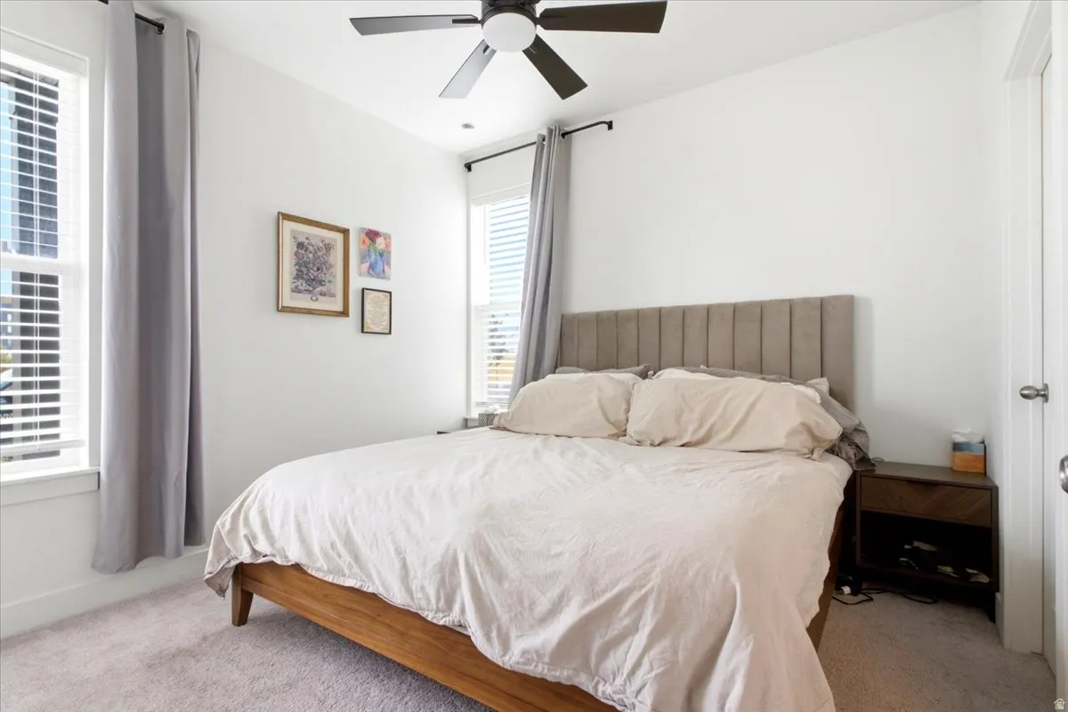 Carpeted bedroom featuring two windows with blinds and drapery, a ceiling fan with integrated lighting, and a white interior door