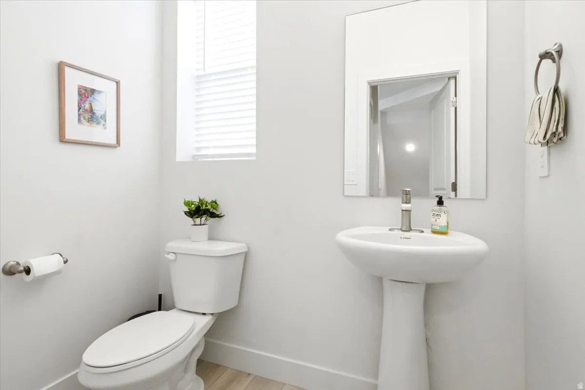 Powder room featuring a pedestal sink with a single-handle faucet, a wall-mounted mirror, a standard toilet, and wood-finish flooring