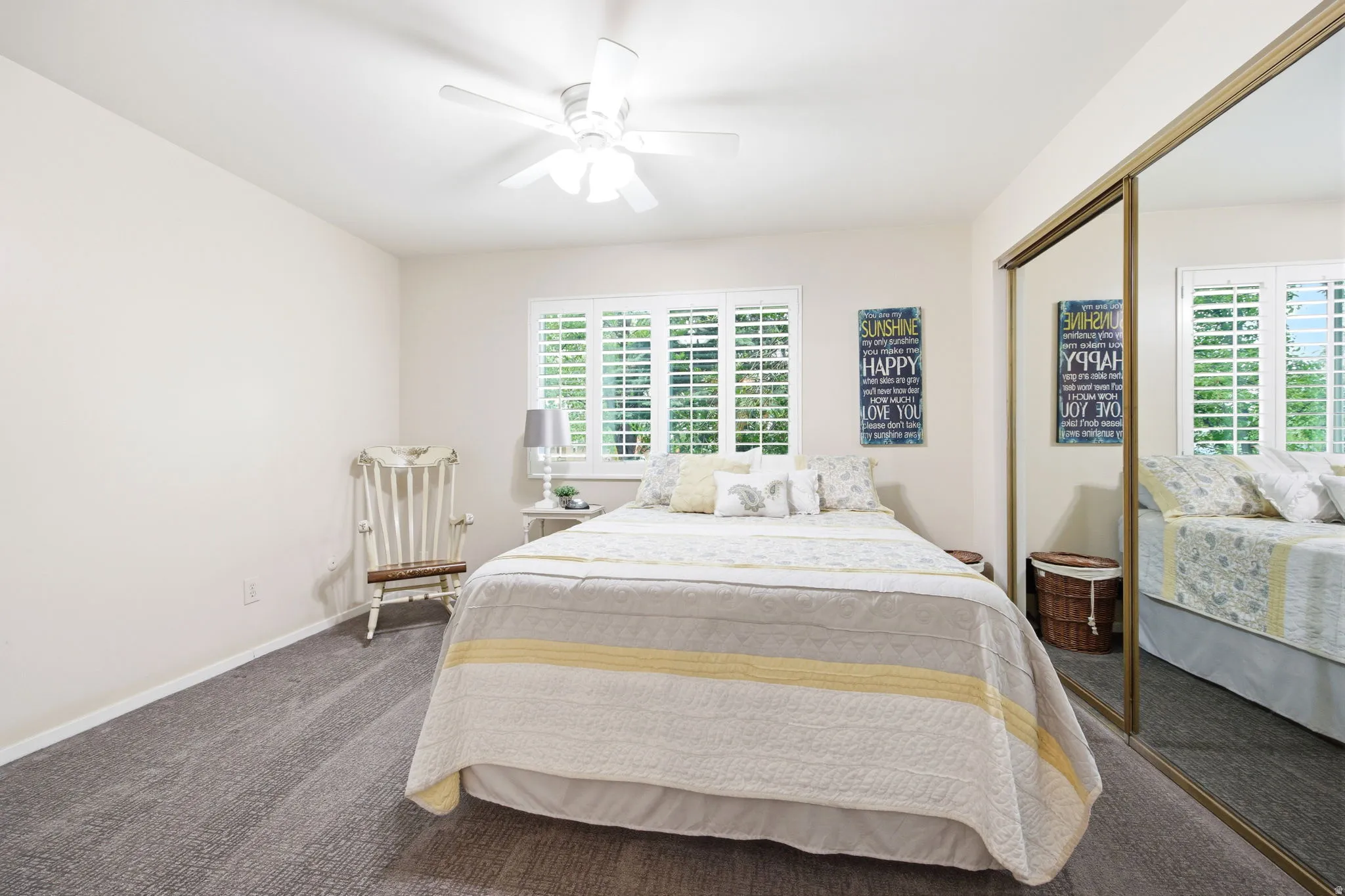 Bedroom featuring white plantation shutters, neutral tone walls, gray carpet flooring, and a ceiling fan