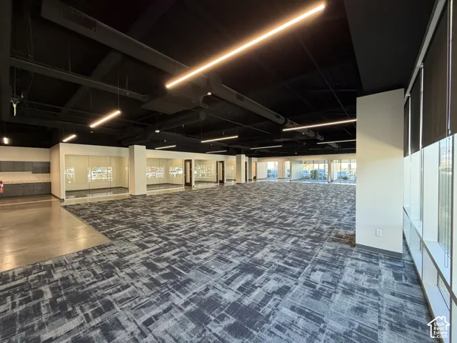 Open layout with patterned carpet, exposed black ceiling, modern linear lighting, and large exterior windows with roller shades